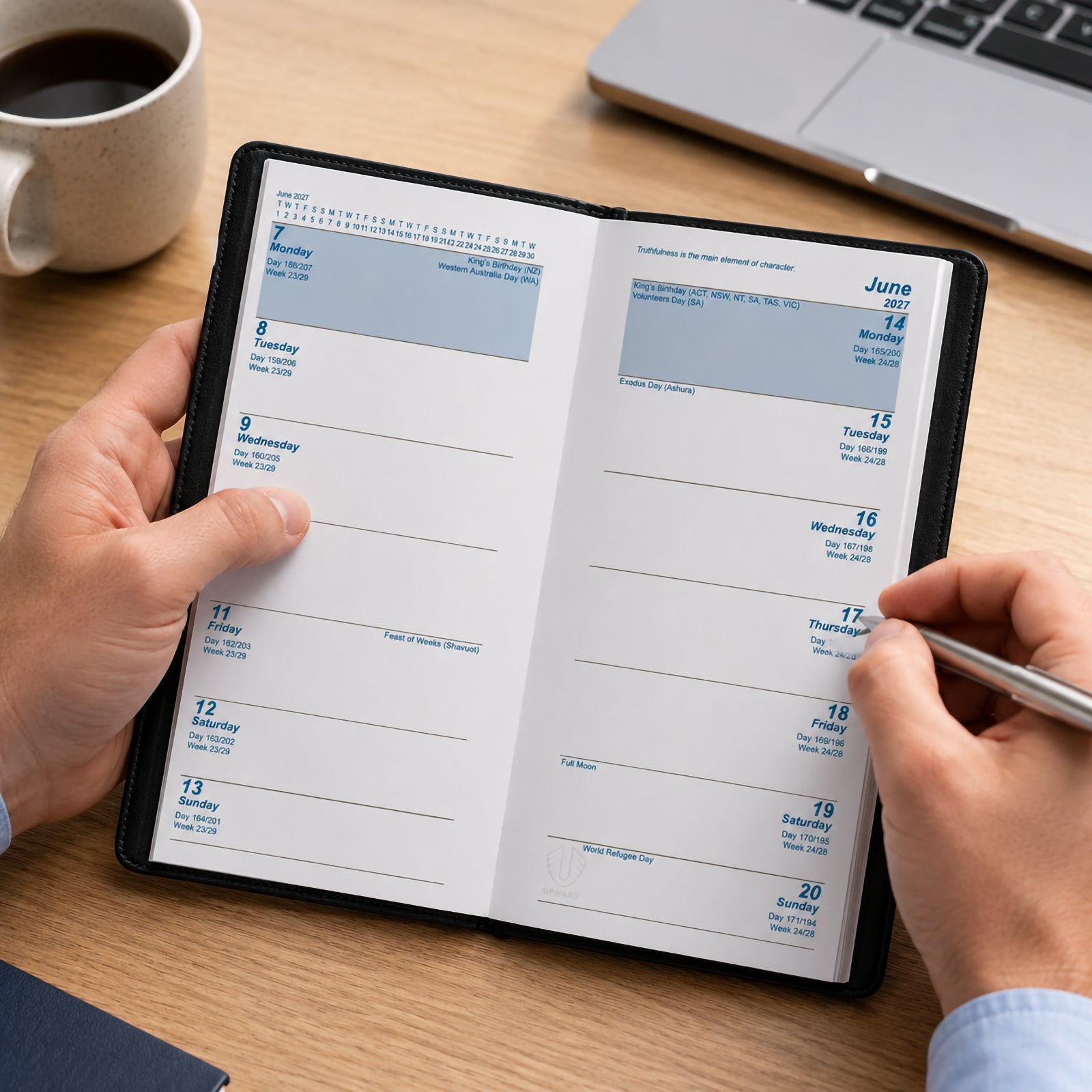 A user writes in the Upward Standard 2027 A6/7 Two Week To View Diary while seated at a desk with a laptop and coffee mug nearby. The open spread displays a clear two week planning layout, providing ample space for appointments, reminders and upcoming commitments in a compact portable format.