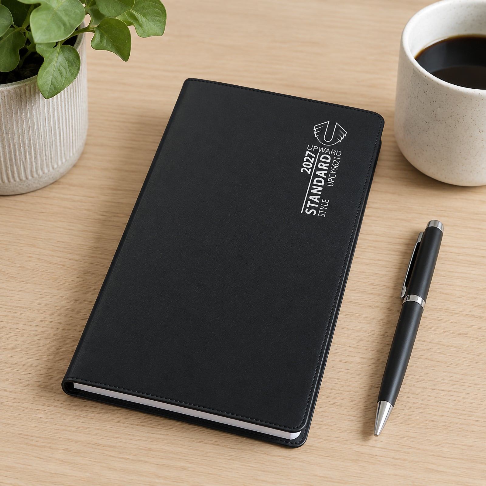 The closed black Upward Standard 2027 diary sits on a timber desk beside a pen, coffee cup and indoor plant. The professional PVC cover and slim portable design make it suitable for carrying daily schedules, appointments and important dates wherever needed.