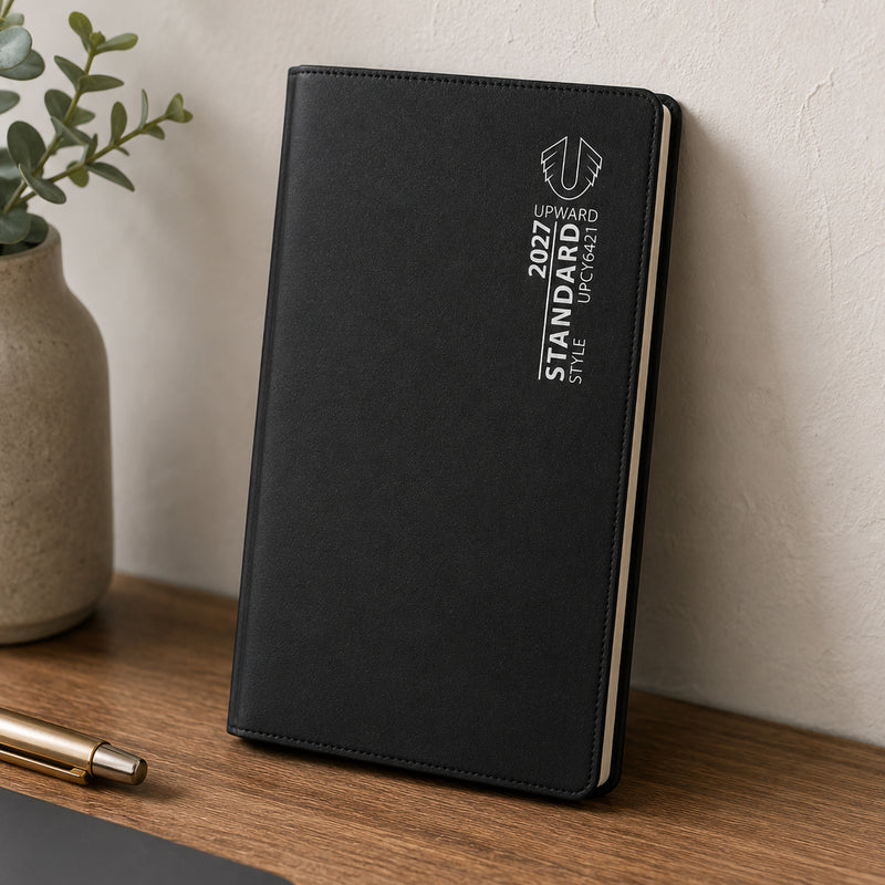 The diary stands upright on a wooden shelf beside decorative greenery and office accessories. The durable black cover and slim profile highlight its suitability for professionals, students and anyone seeking reliable weekly scheduling throughout 2027.