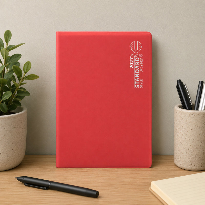 The diary stands upright on a tidy workspace beside stationery and a decorative plant. The bright red cover creates a bold professional statement while offering a practical solution for managing weekly commitments throughout the year.
