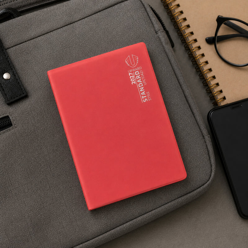 The red Upward Standard 2027 diary rests on top of a laptop bag alongside a smartphone, notebook and glasses. Its compact A6 format highlights its convenience for professionals, students and travellers who need a reliable weekly planner on the move.