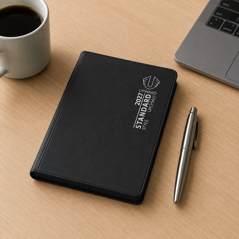 The closed A7 diary rests on a desktop workspace next to a pen, laptop and coffee mug. Its durable black PVC cover and compact format demonstrate a practical planning solution for professionals, students and everyday organisation.