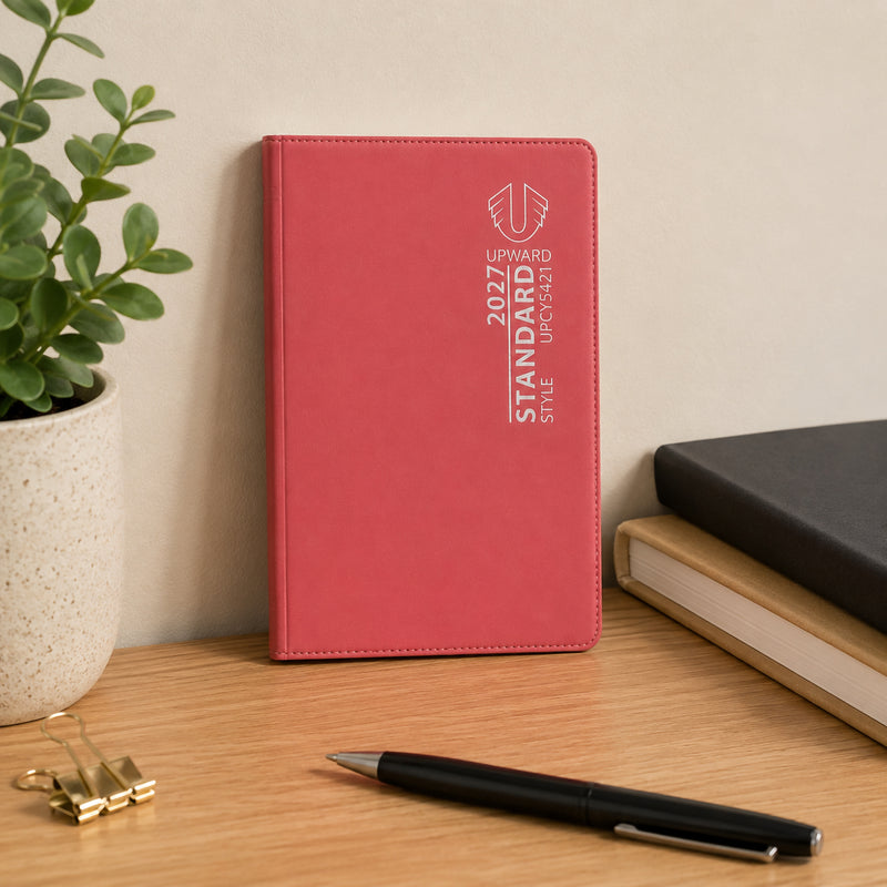 The diary stands upright on a desk beside office accessories and a potted plant. Its bold red cover creates strong visual appeal while showcasing a practical planning tool designed to help users organise tasks, meetings and important dates throughout 2027.