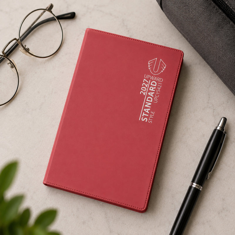 The red Upward Standard 2027 A7 diary is displayed on a marble surface with reading glasses, a pen and a travel bag. The image emphasises its compact design and convenience for professionals, students and travellers who need quick access to weekly planning information.