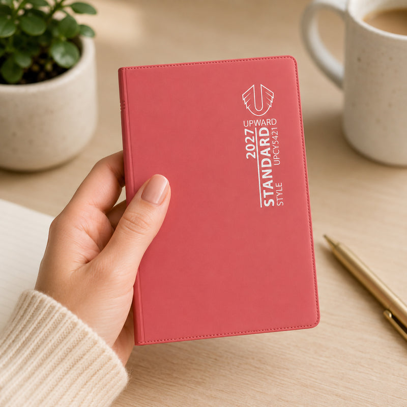 A person holds the Upward Standard 2027 A7 Week To View Diary featuring a vibrant red cover in a bright workspace setting. The compact pocket sized diary is shown alongside a coffee mug and notebook, highlighting its portability and suitability for managing appointments, reminders and weekly schedules on the go.