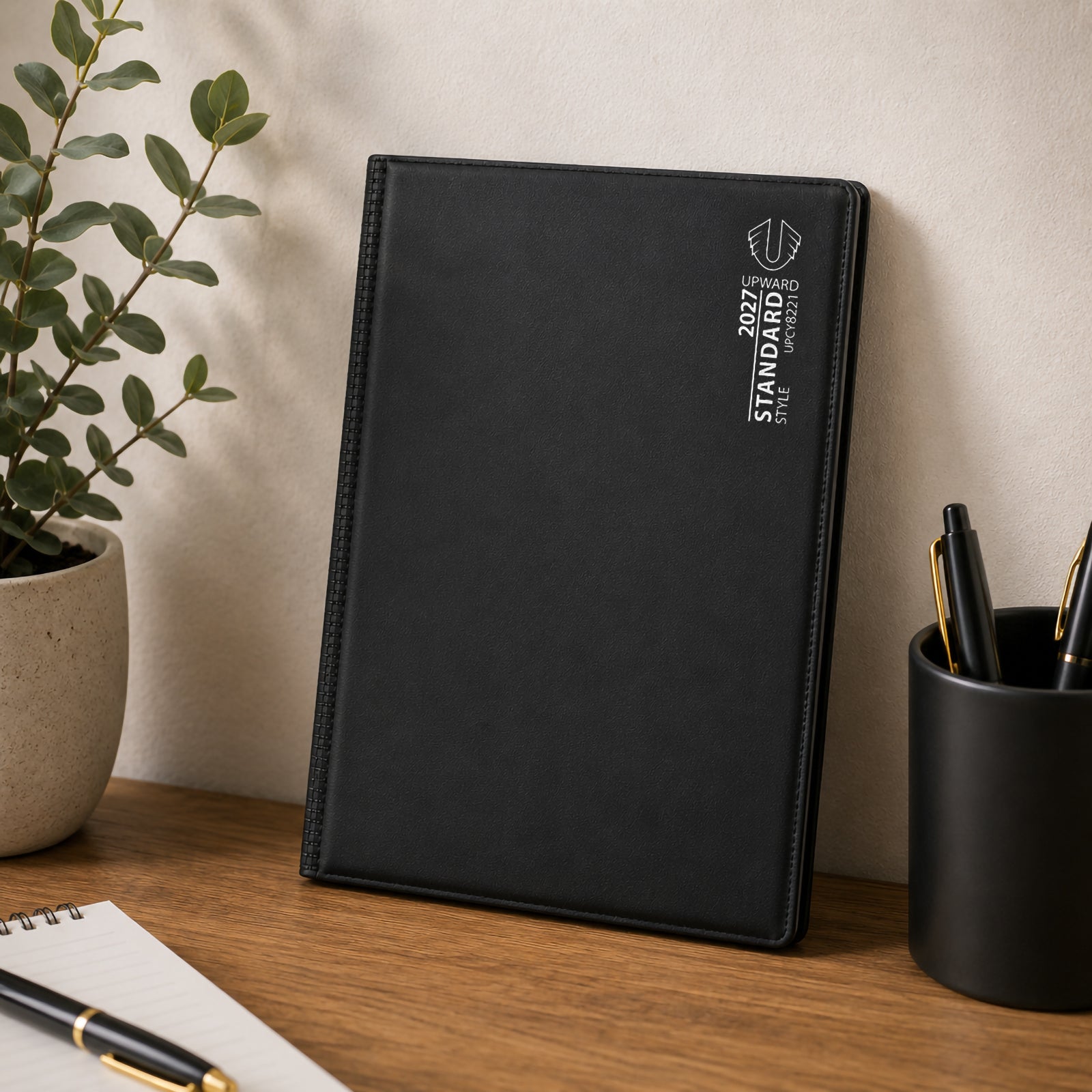 Upright view of the diary positioned on a desk with stationery and indoor greenery. The sleek black design and sturdy construction highlight a reliable planning companion for managing schedules, recording notes and tracking goals across 2027.