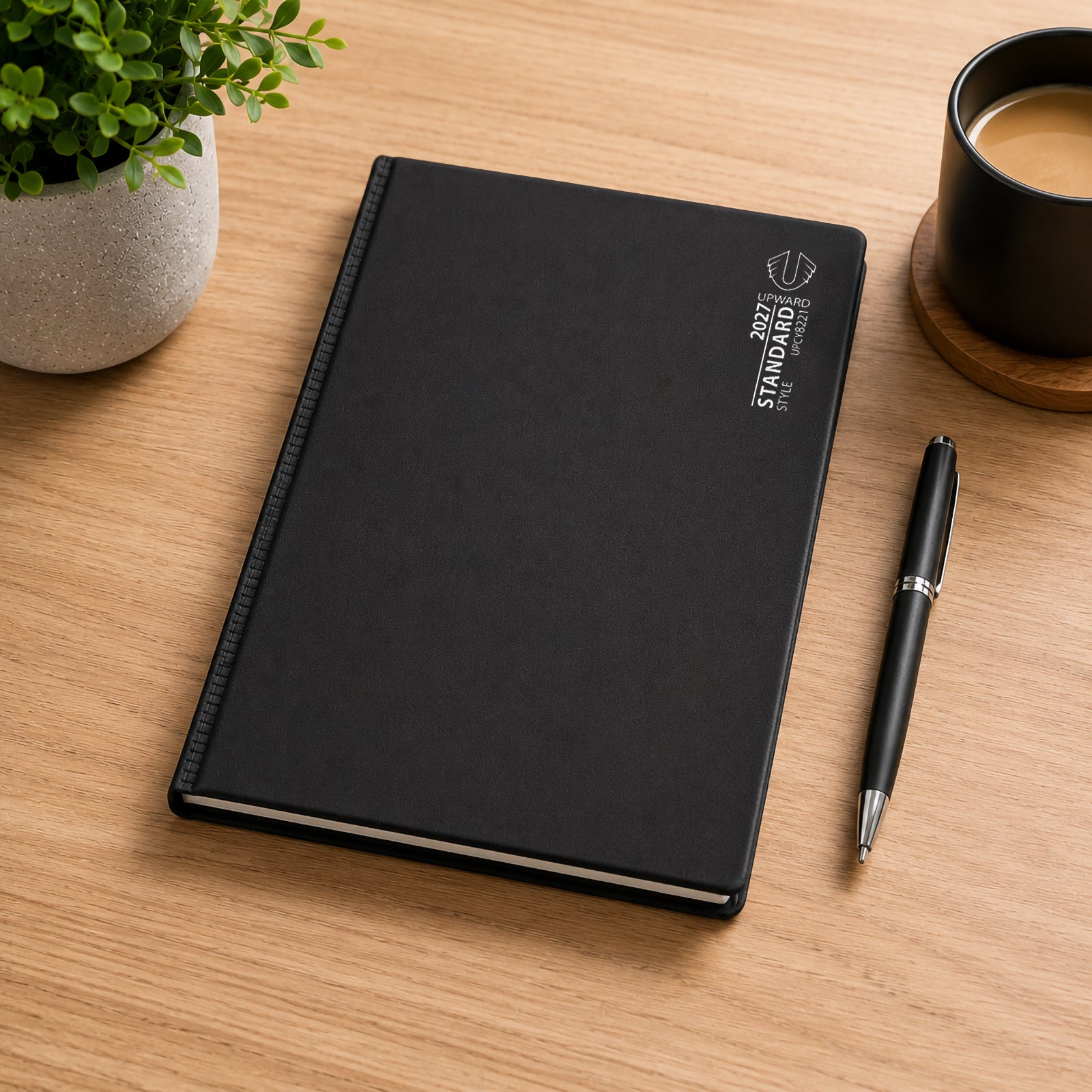 The Upward Standard 2027 diary displayed on a timber desk alongside a pen, coffee cup and decorative plant. The durable black PVC cover presents a clean and professional appearance suitable for office workers, students and anyone who values structured daily planning.