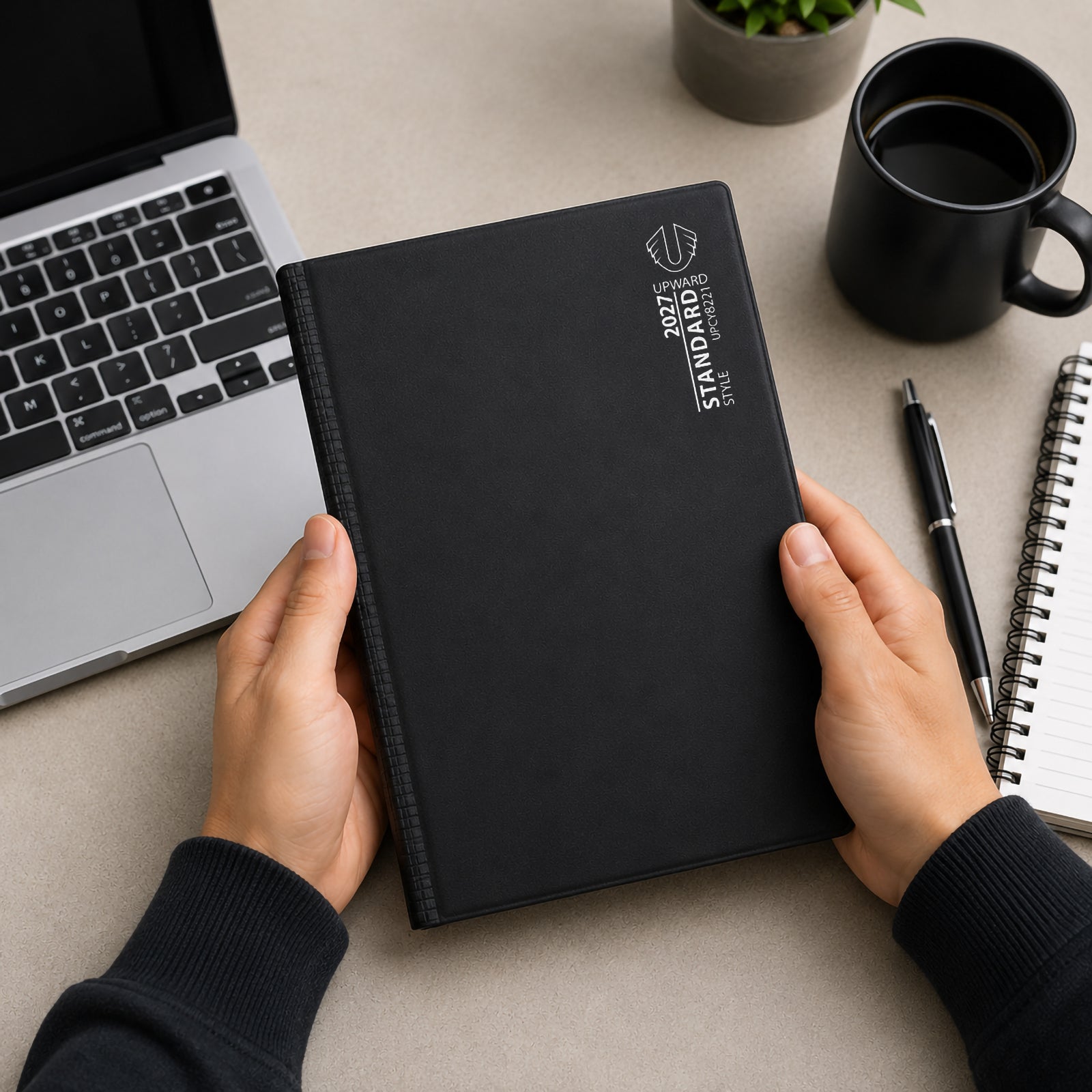 A user holds the Upward Standard 2027 B5 Day To Page Diary at a modern workstation beside a laptop, notebook and coffee mug. The professional black cover and practical B5 size demonstrate its suitability for organising appointments, meetings, daily tasks and long term planning throughout the year.