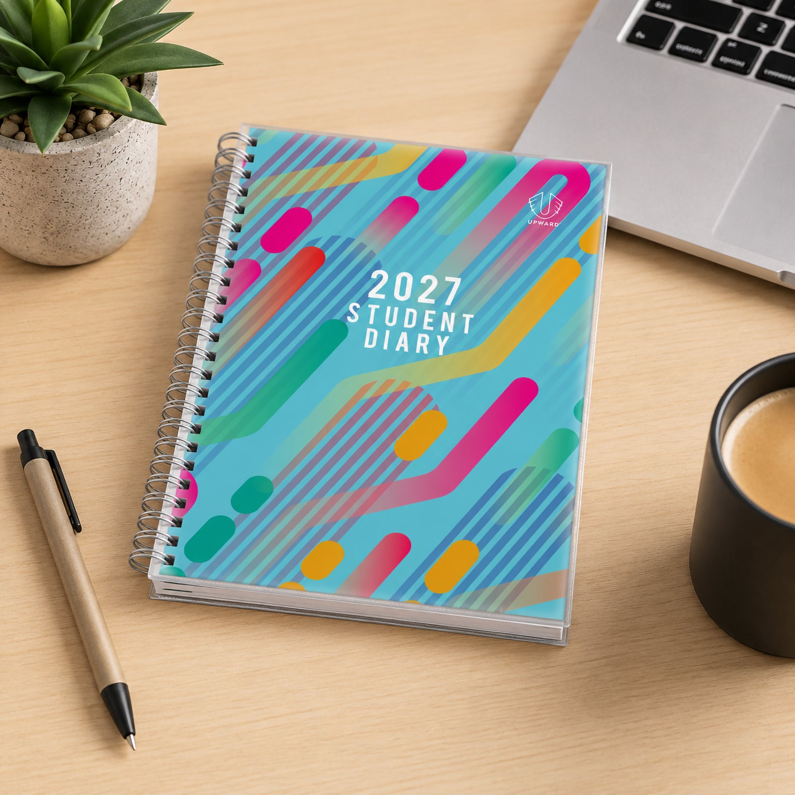 The Upward Student 2027 diary displayed on a study desk beside a laptop, coffee cup and indoor plant. The colourful cover design creates a modern academic look while demonstrating its suitability for home study, schoolwork and personal organisation.