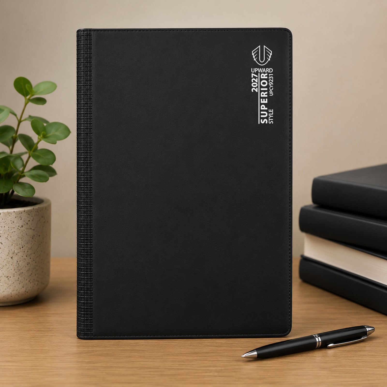 Front facing product display of the Upward Superior A4 diary positioned on a desk alongside stacked books and a pen. The durable black cover and textured spine reinforce its professional appearance and everyday reliability.