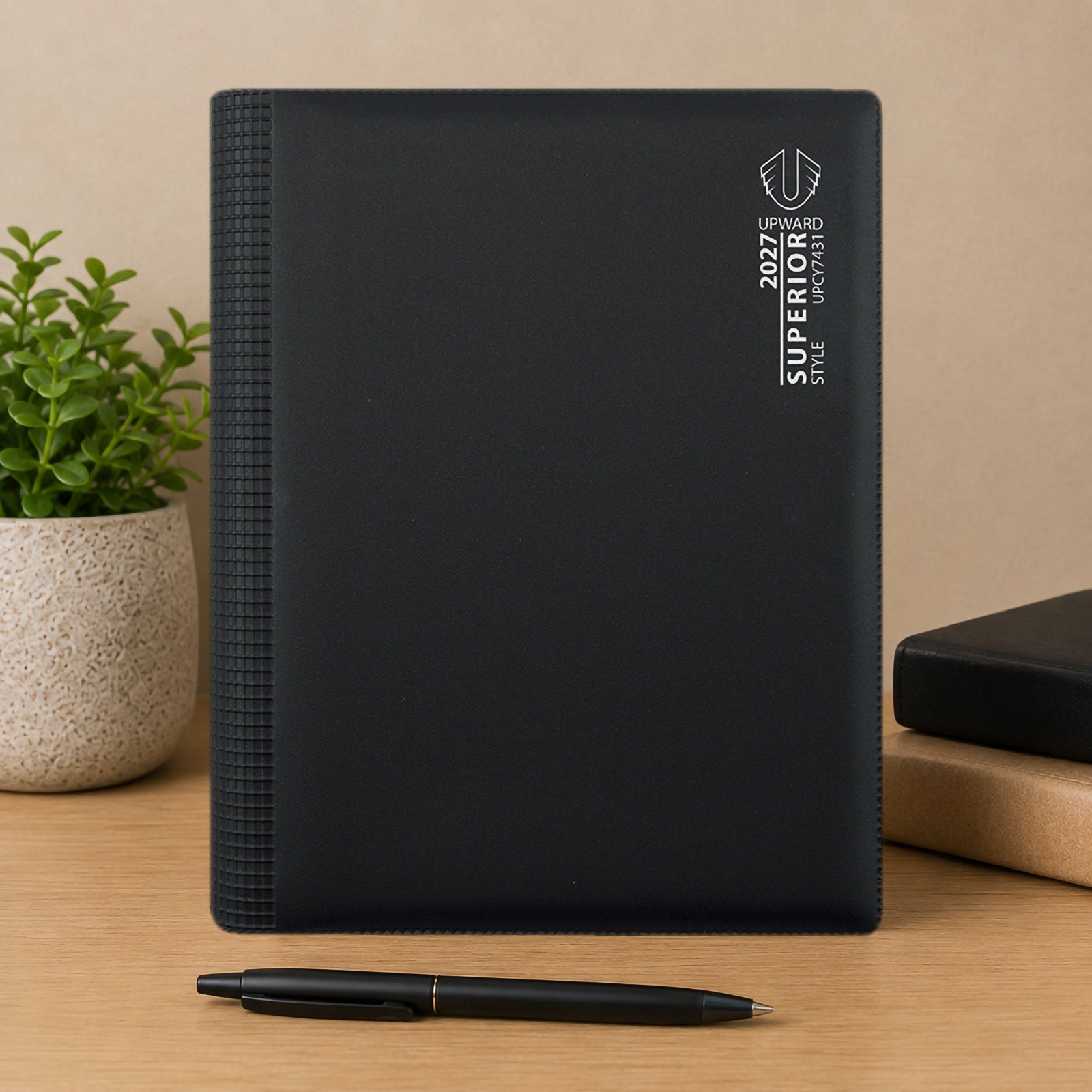 Front cover product display featuring the Upward Superior 2027 A5 diary standing upright on a desk alongside books and a decorative plant. The clean black finish and professional branding create a polished appearance suitable for workplace use.
