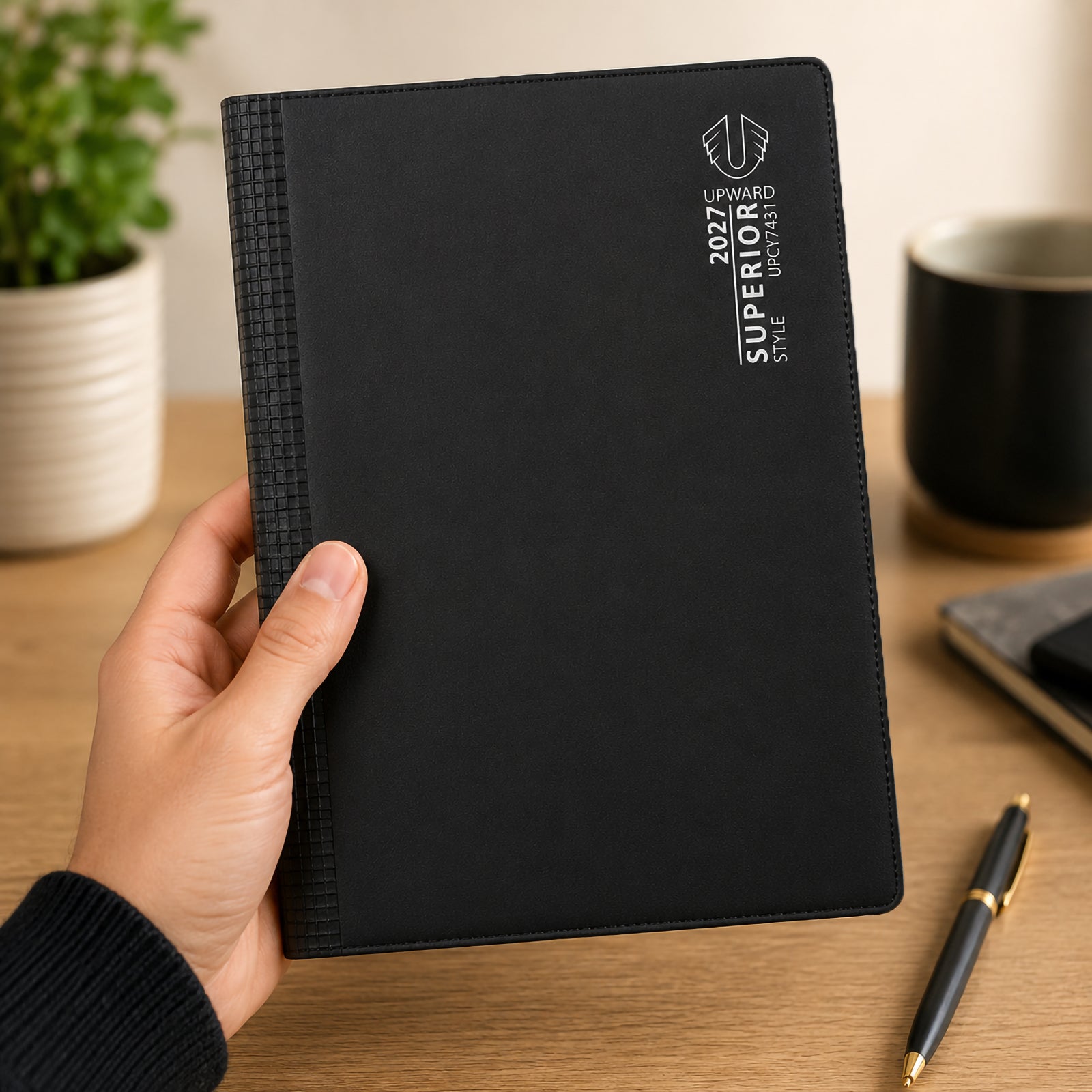 Hand holding the Upward Superior 2027 A5 Week To View Diary showcasing its professional black PVC cover and practical A5 size. The diary is displayed in a modern workspace environment, highlighting portability, durability and suitability for everyday planning.