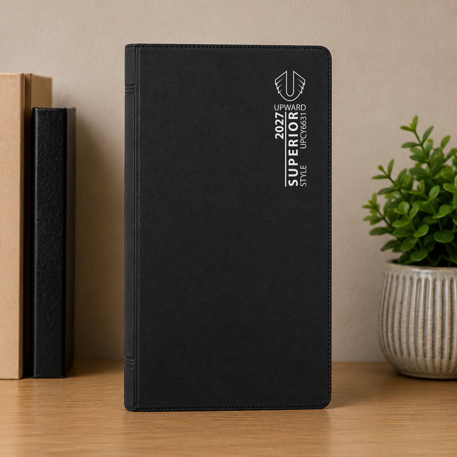 Upright front cover view of the Upward Superior 2027 A6/7 diary displayed on a desk with books and a decorative plant. The professional black finish presents a clean and organised appearance suitable for office environments.