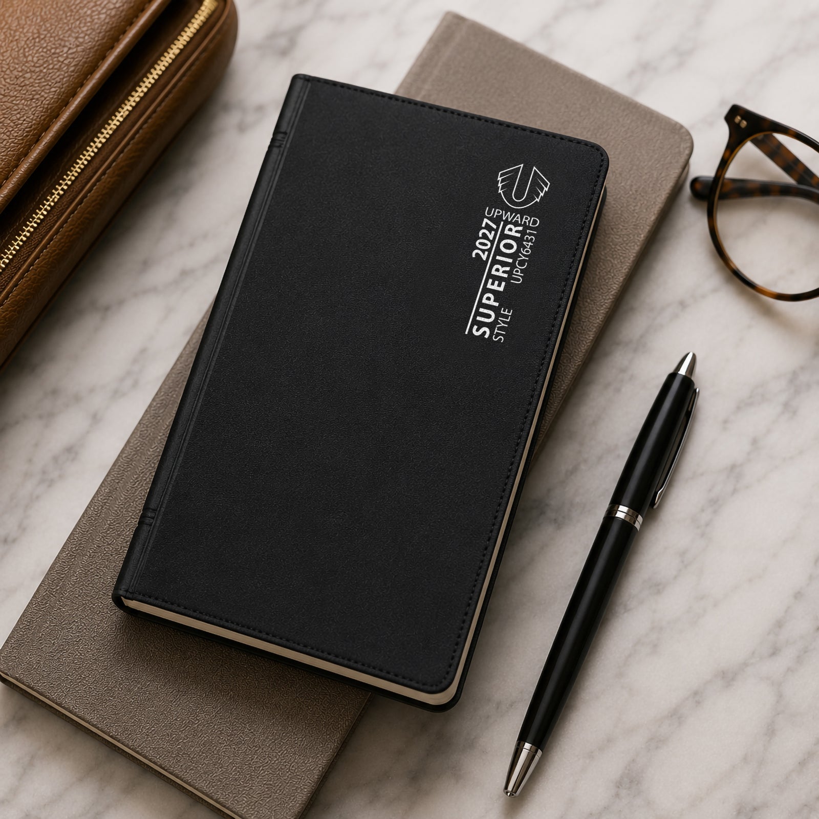 Closed Upward Superior 2027 A6/7 diary resting on top of a notebook alongside a pen, glasses and travel accessories. The image highlights the professional black PVC cover and convenient portable size suitable for work, study and travel.