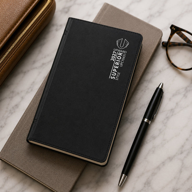 Closed Upward Superior 2027 A6/7 diary resting on top of a notebook alongside a pen, glasses and travel accessories. The image highlights the professional black PVC cover and convenient portable size suitable for work, study and travel.