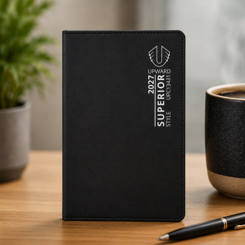 Front standing view of the Upward Superior 2027 A7 Week To View Diary in black. The sleek cover design and compact dimensions are showcased in a professional workspace setting, demonstrating its suitability for portable planning and organisation.