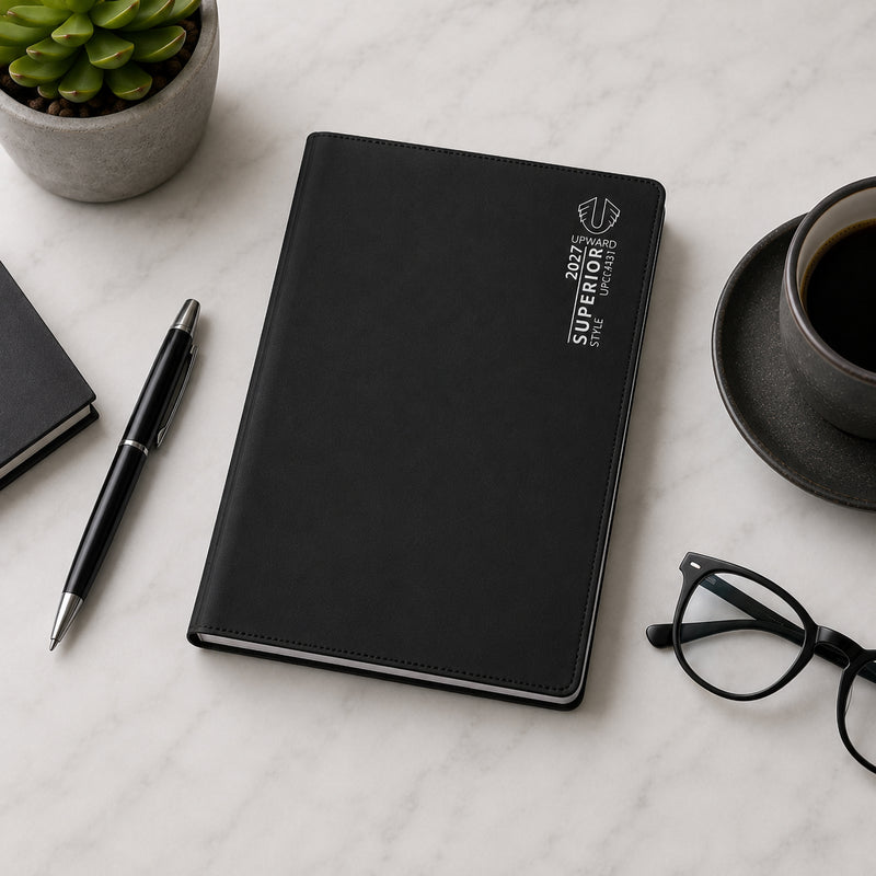 Closed Upward Superior 2027 A7 diary with black PVC cover positioned on a marble surface alongside office accessories including a pen, coffee cup and reading glasses. The image highlights the compact size and professional appearance suitable for everyday business use.