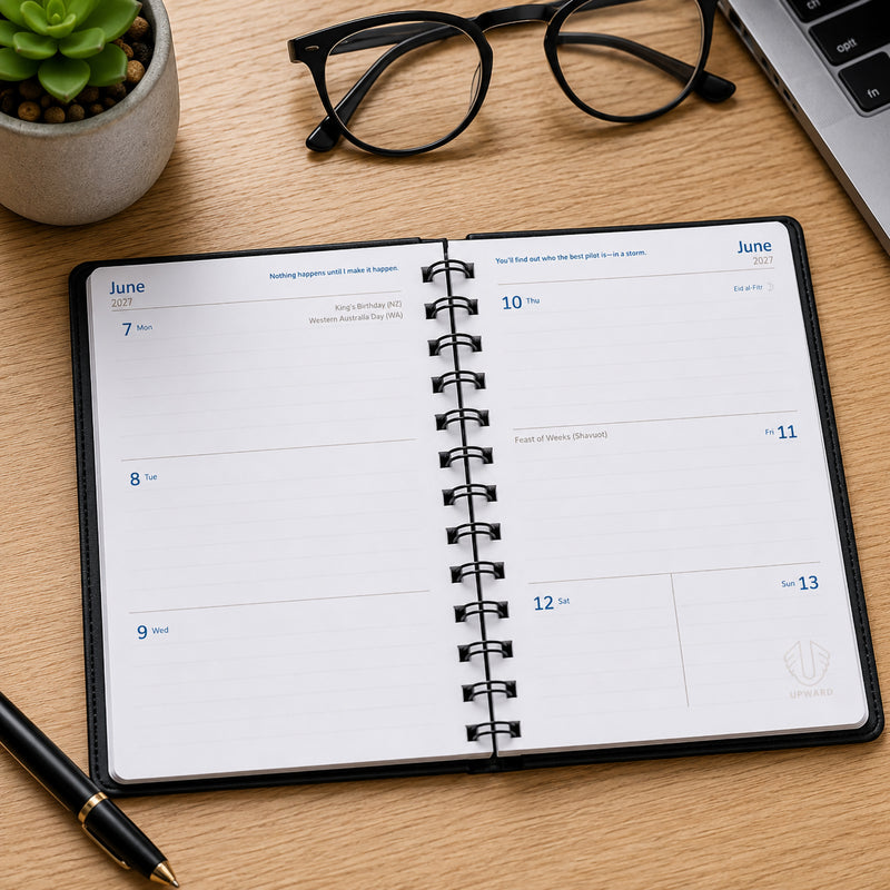 Open Upward Superior 2027 A7 Week To View Diary displayed on a timber desk beside glasses, a laptop and a pen. The compact weekly planner shows a clear week to view layout with dedicated space for appointments, reminders and daily scheduling in a highly portable format.