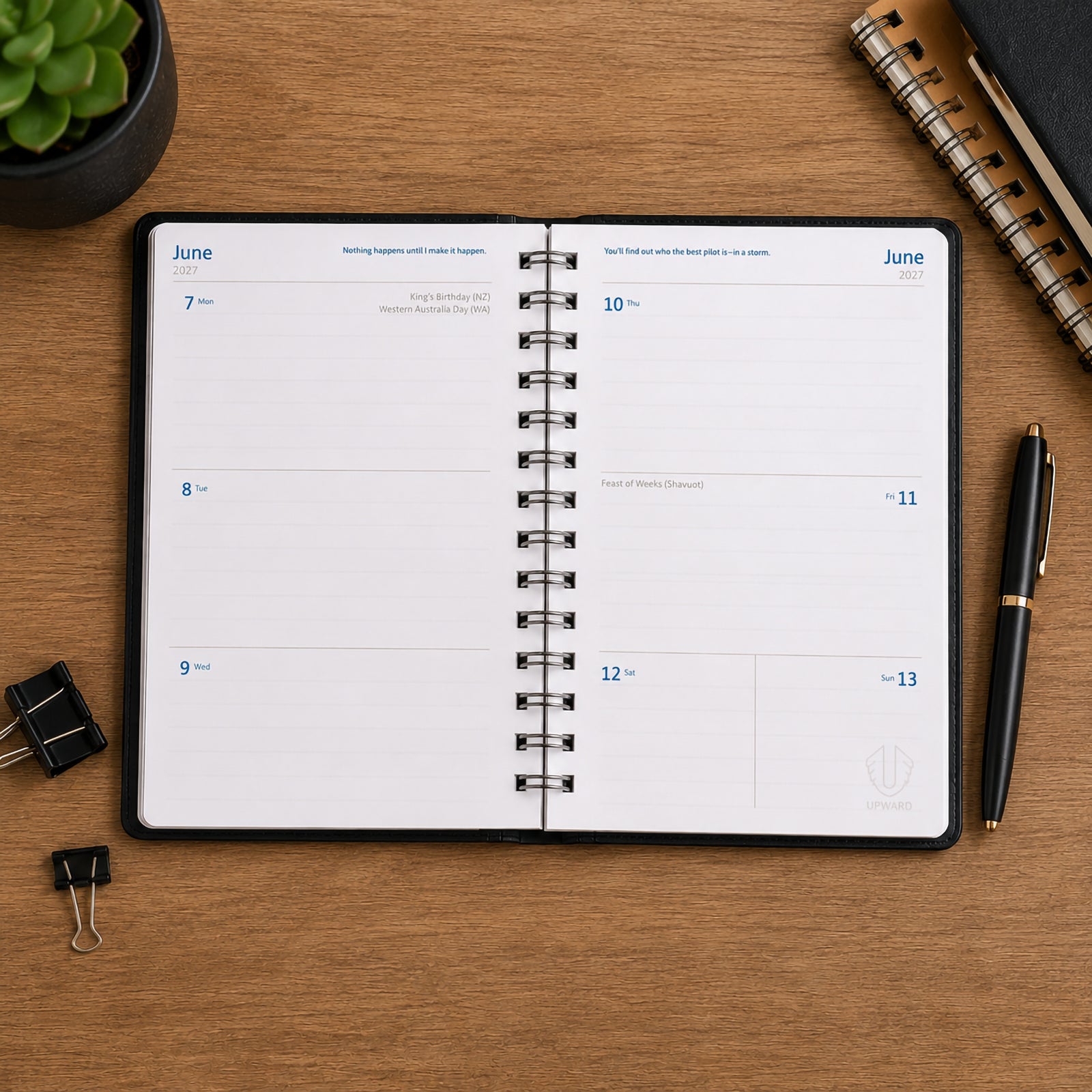 Open view of the Upward Superior 2027 B6 Week To View Diary with black cover displayed on a timber desk. The weekly layout provides generous writing space for appointments, meetings, reminders and task management, with stationery accessories positioned around the diary to create a professional planning workspace.