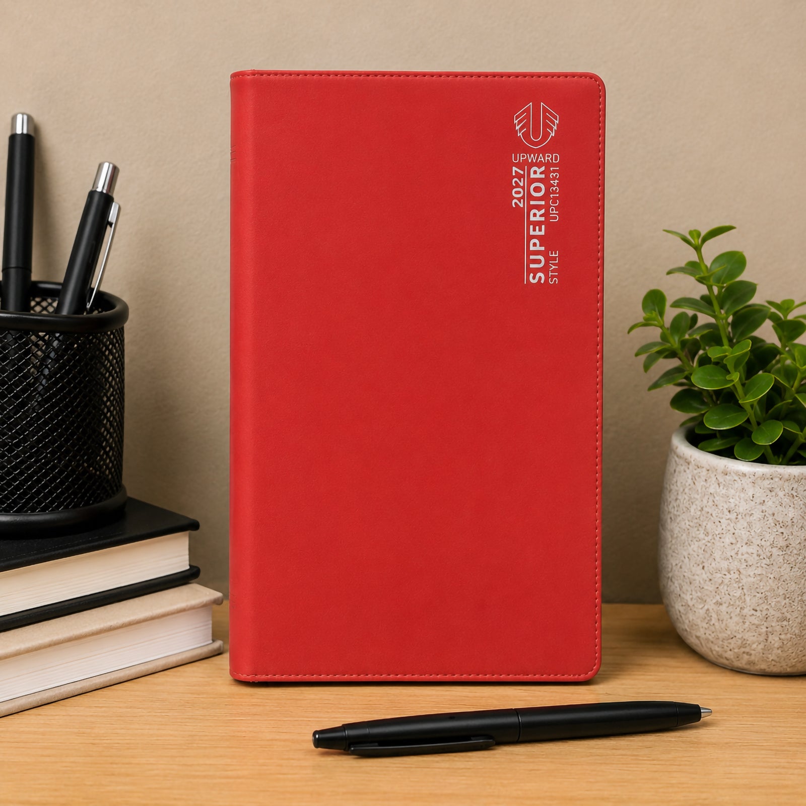 Front standing view of the Upward Superior 2027 B6 Week To View Diary in red. Positioned beside books, pens and a potted plant, the diary highlights its durable cover, compact design and suitability for office, study and home planning.