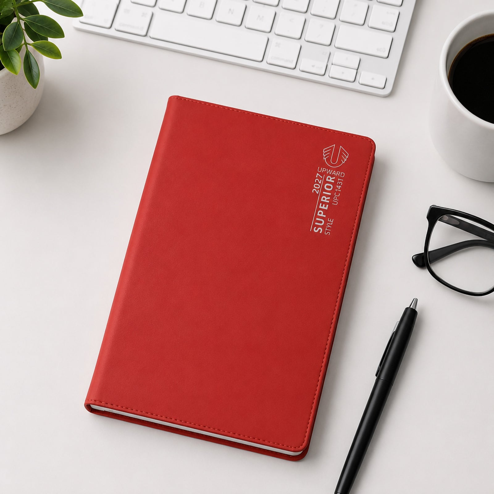 The Upward Superior 2027 B6 diary with a vibrant red PVC cover is displayed on a modern desktop alongside a keyboard, coffee cup, pen and glasses. The compact size and professional appearance make it ideal for portable planning and daily organisation.