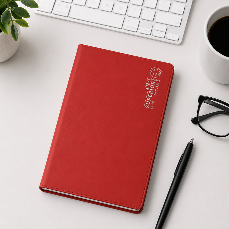 The Upward Superior 2027 B6 diary with a vibrant red PVC cover is displayed on a modern desktop alongside a keyboard, coffee cup, pen and glasses. The compact size and professional appearance make it ideal for portable planning and daily organisation.