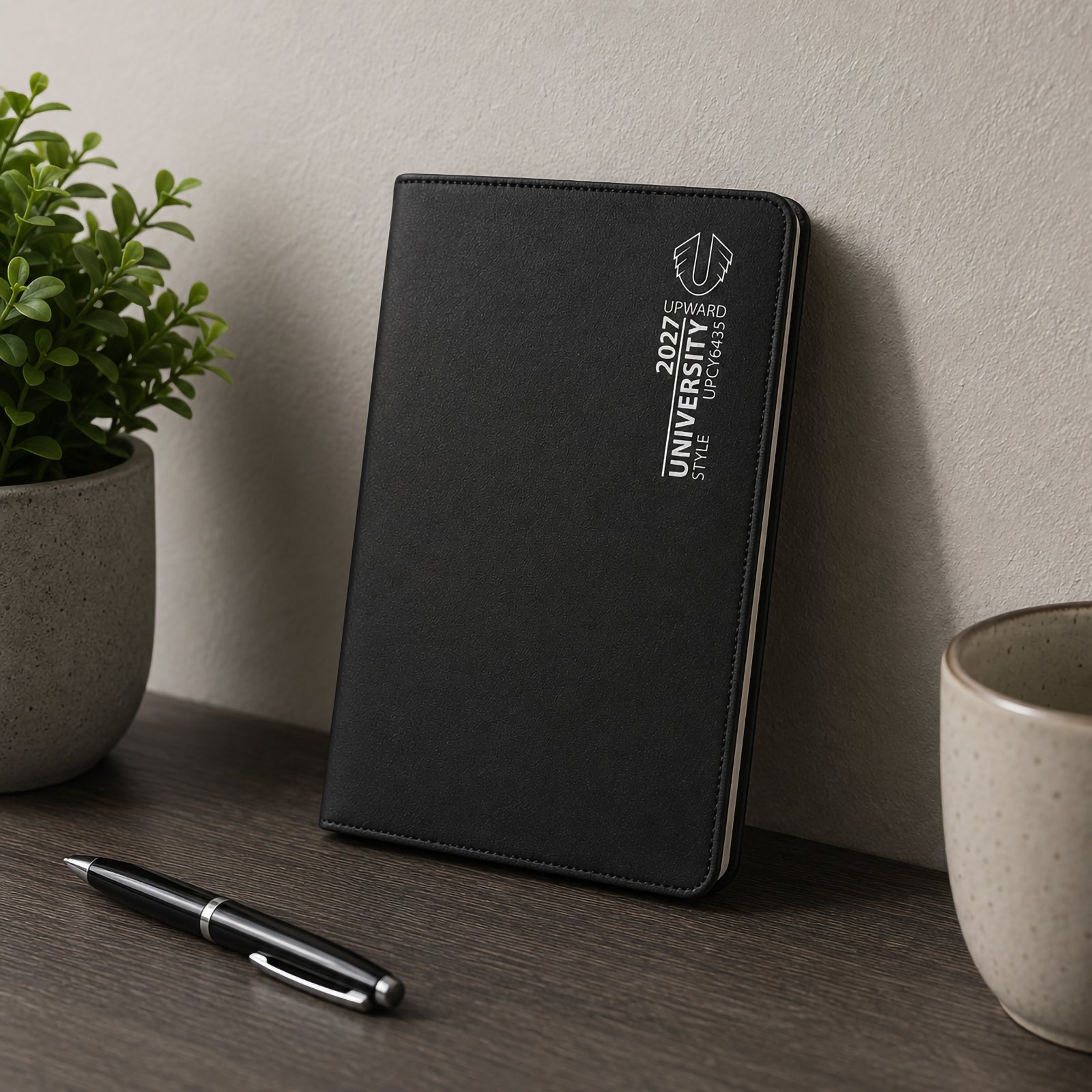 The Upward University 2027 diary is displayed upright on a desk alongside stationery and indoor plants. The professional black cover and compact format make it suitable for managing university schedules, appointments, deadlines and weekly planning.