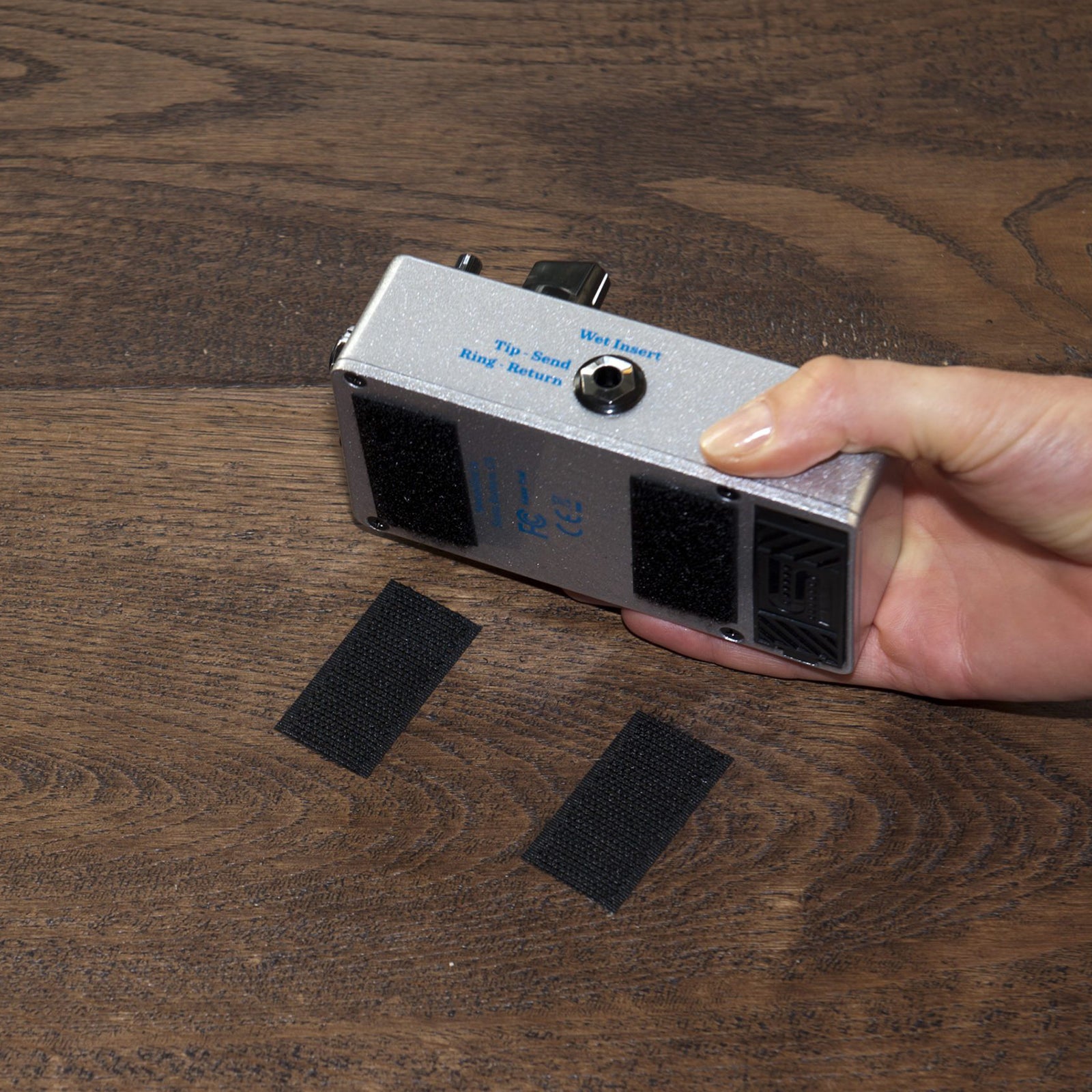 A hand holds a compact electronic device fitted with black heavy duty Velcro strips above a wooden surface. The image demonstrates secure removable fastening for equipment, electronics and workshop accessories.