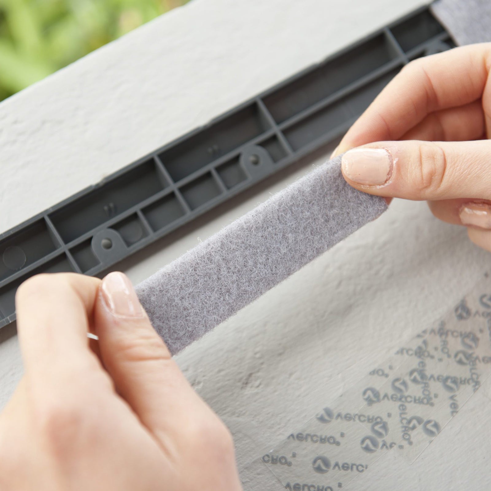 Close up image of hands applying grey VELCRO Brand heavy duty hook and loop strip onto a rough mounting surface for secure indoor fastening.