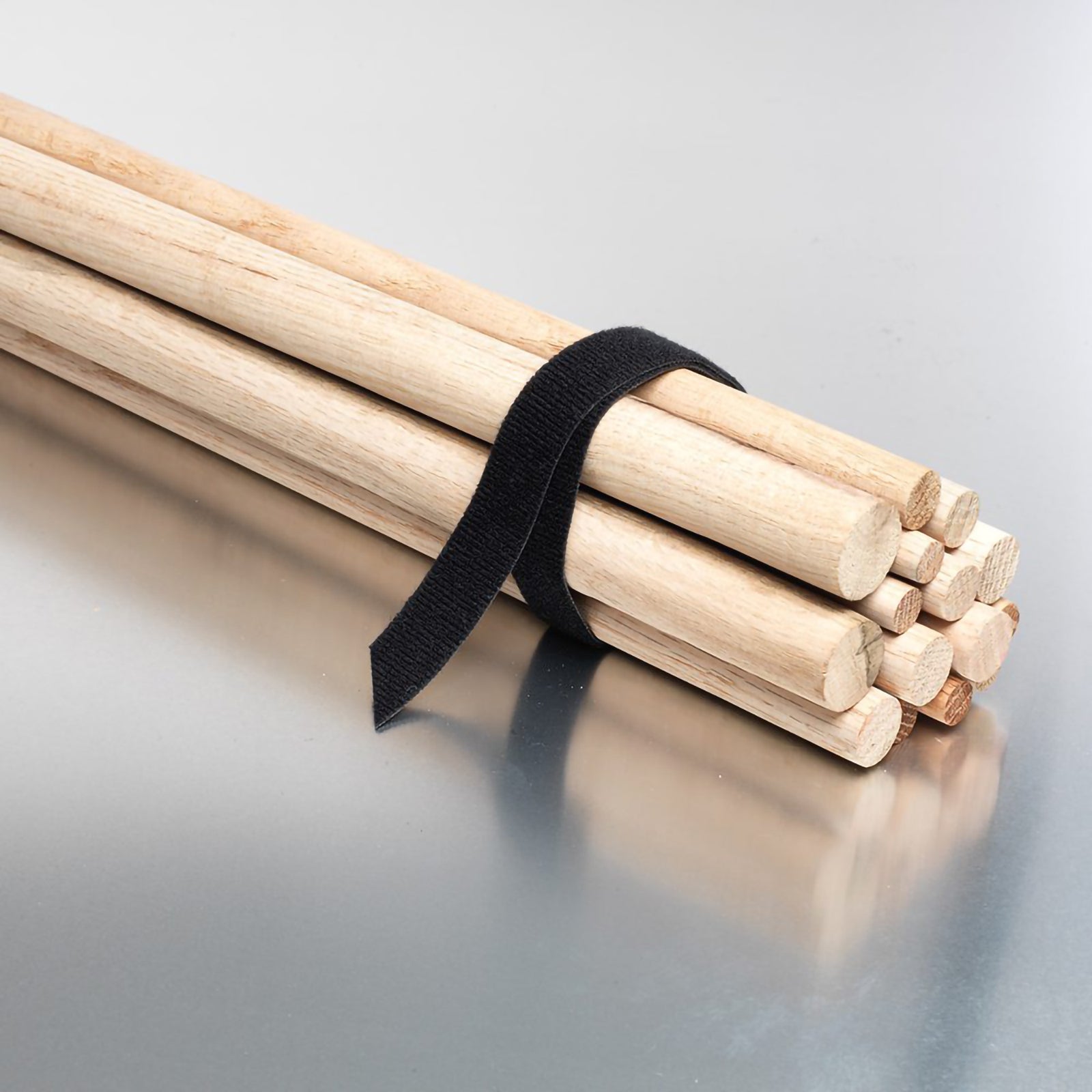 Black reusable Velcro bundling strap wrapped around a stack of wooden poles on a reflective surface. The image demonstrates secure fastening strength and adjustable bundling for tools, timber and workshop materials.