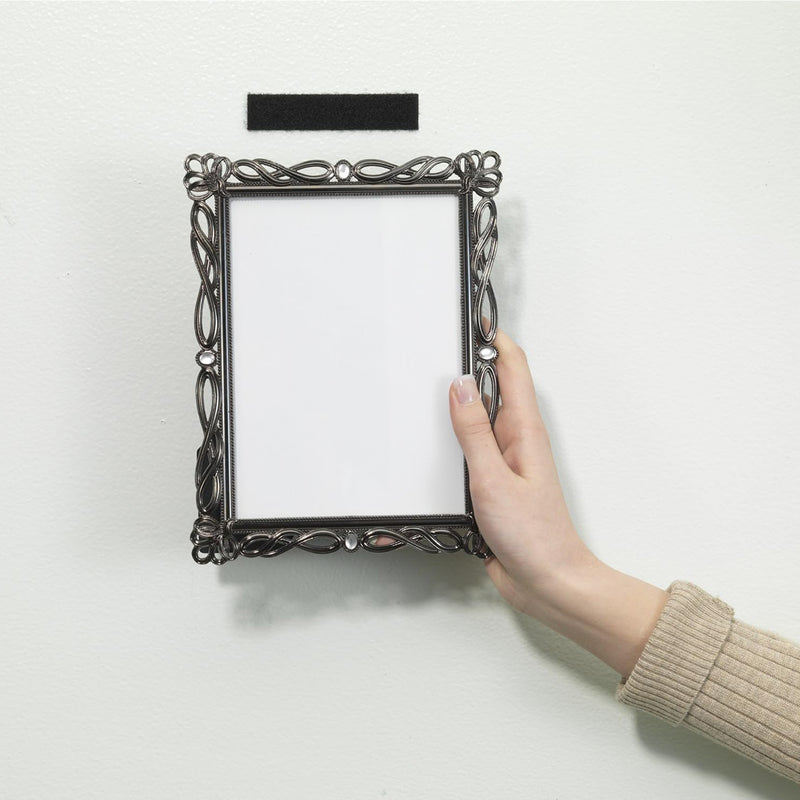 A decorative black picture frame is being positioned against a wall using black Velcro stick on tape. The image demonstrates simple removable mounting for lightweight home decor and office display applications.