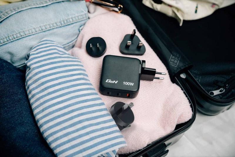 Verbatim Mini GaN Charger 100W 30242 packed in an open suitcase beside clothing with its interchangeable travel plug heads