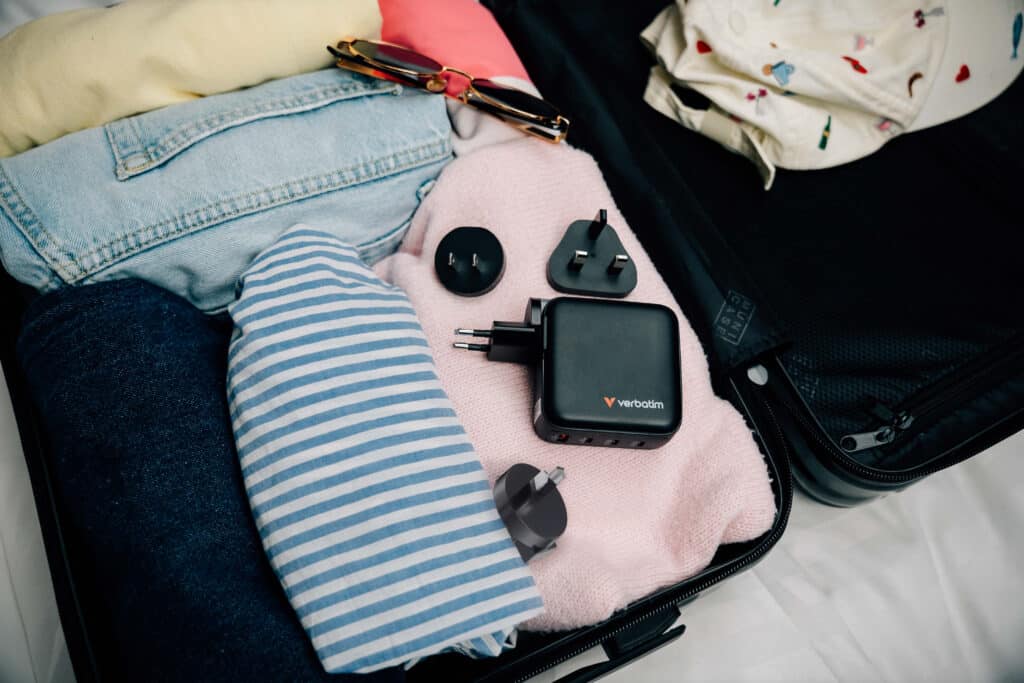 Verbatim Mini GaN Charger 140W 30243 packed in an open suitcase beside clothing with its interchangeable travel plug heads