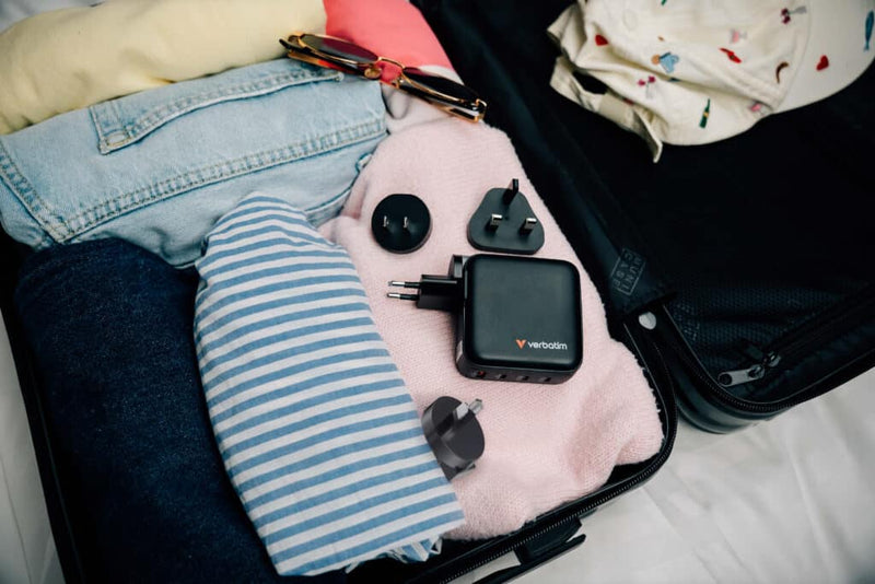 Verbatim Mini GaN Charger 140W 30243 packed in an open suitcase beside clothing with its interchangeable travel plug heads
