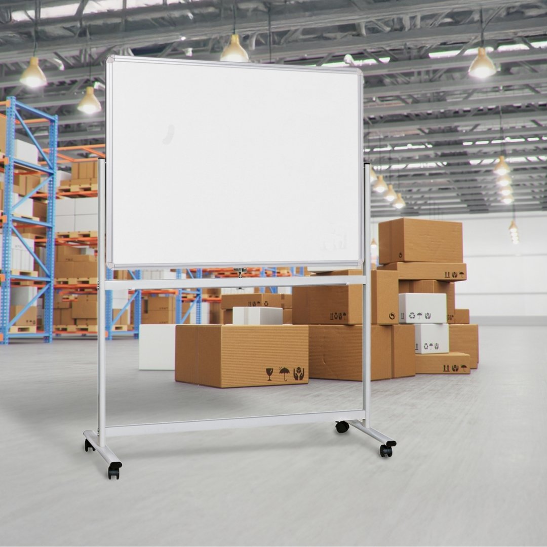 The Chilli mobile whiteboard standing on a concrete warehouse floor with cardboard cartons and steel racking behind it, showing the board at working height on its wheeled silver frame.