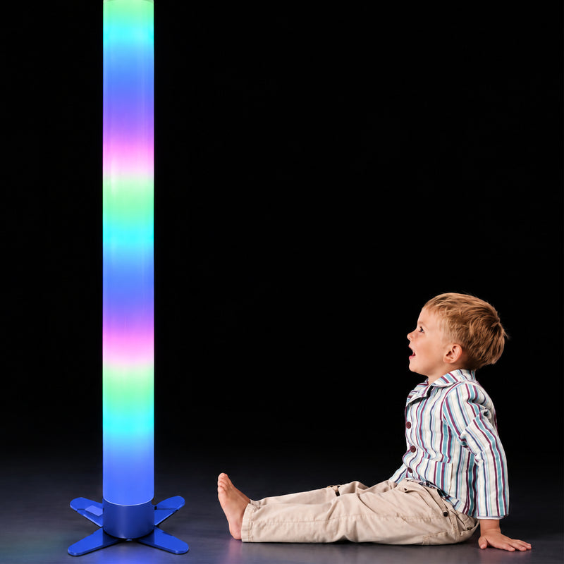 Child seated beside the MTA Essentials Waterless LED Tube while watching the tall illuminated column. Bands of green, blue and pink light travel through the 183cm tube against a dark background, highlighting its strong visual effect and suitability for supervised observation, sensory engagement and colour exploration.