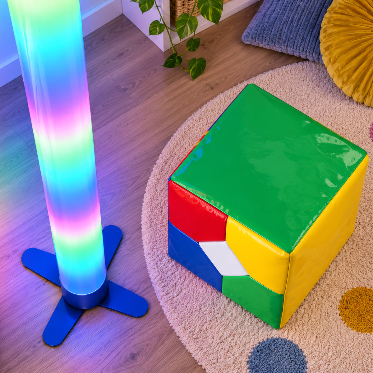 Top view of the MTA Essentials Waterless LED Tube and Extra Large Remote Cube arranged in a sensory space. The illuminated tube stands on its supportive base beside the colourful padded cube, clearly showing the green top and multicoloured sides while soft furnishings and a textured rug create an inviting environment for sensory activities.