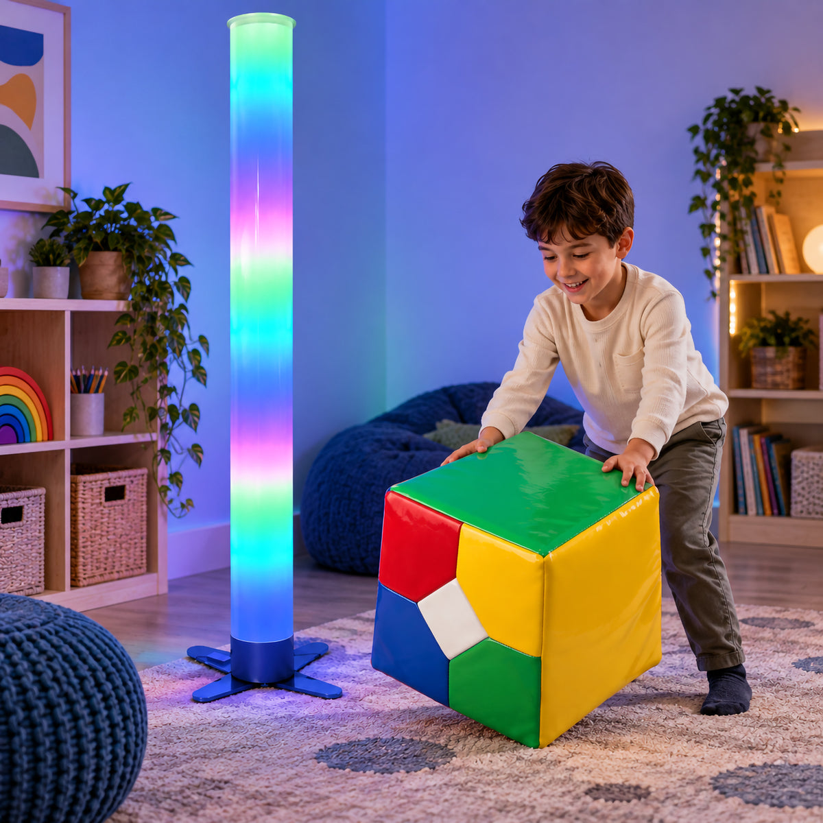 MTA Essentials Waterless LED Tube with Extra Large Remote Cube in a colourful sensory room while a child rolls and explores the large padded cube. The 183cm tube displays bands of blue, green, pink and purple light, demonstrating an engaging interactive setup for supervised sensory exploration, movement and cause and effect activities.
