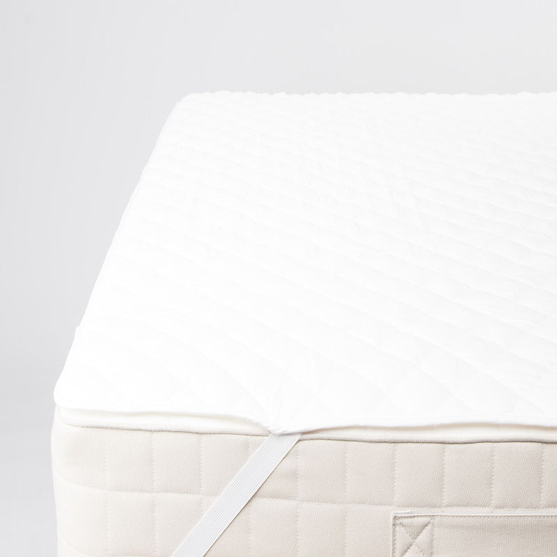 Corner of a mattress with the quilted white protector on top and the white elastic strap pulled down over the corner