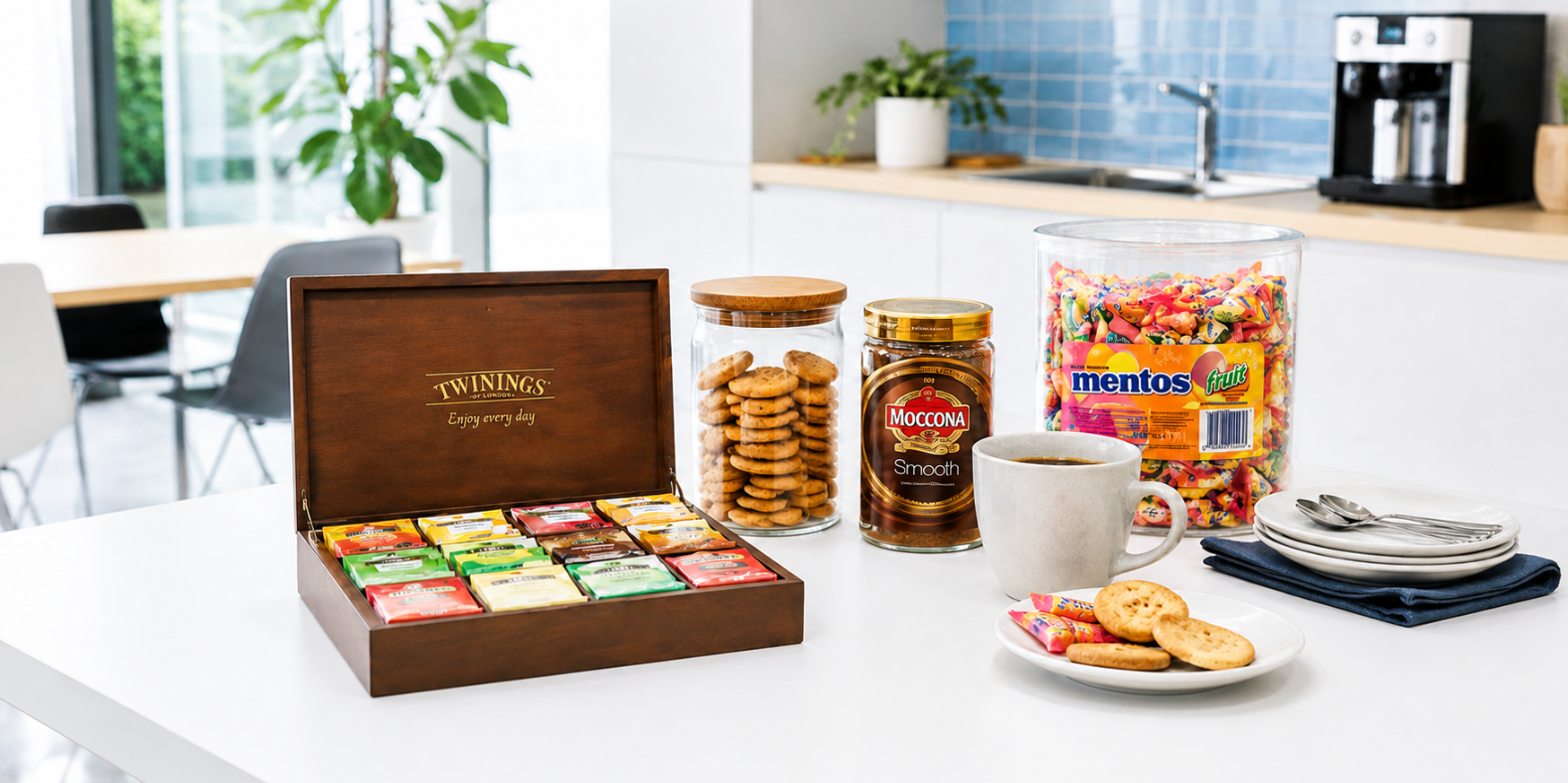 A Twinings tea gift set displayed on a white kitchen counter, featuring an open wooden box of assorted tea packets, a jar of Macgregor Smooth tea, a large clear jar of Mentos candy, and a white mug of tea, with cookies on a plate nearby.