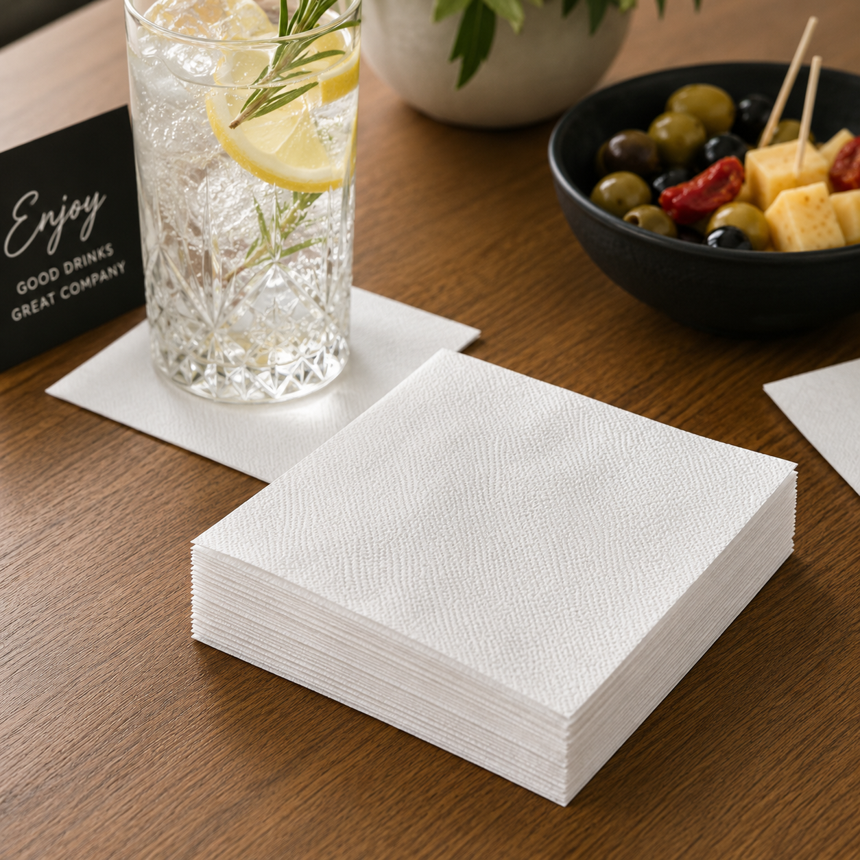 White cocktail napkins stacked on a wooden drinks table beside a glass with lemon, rosemary and shared snacks.