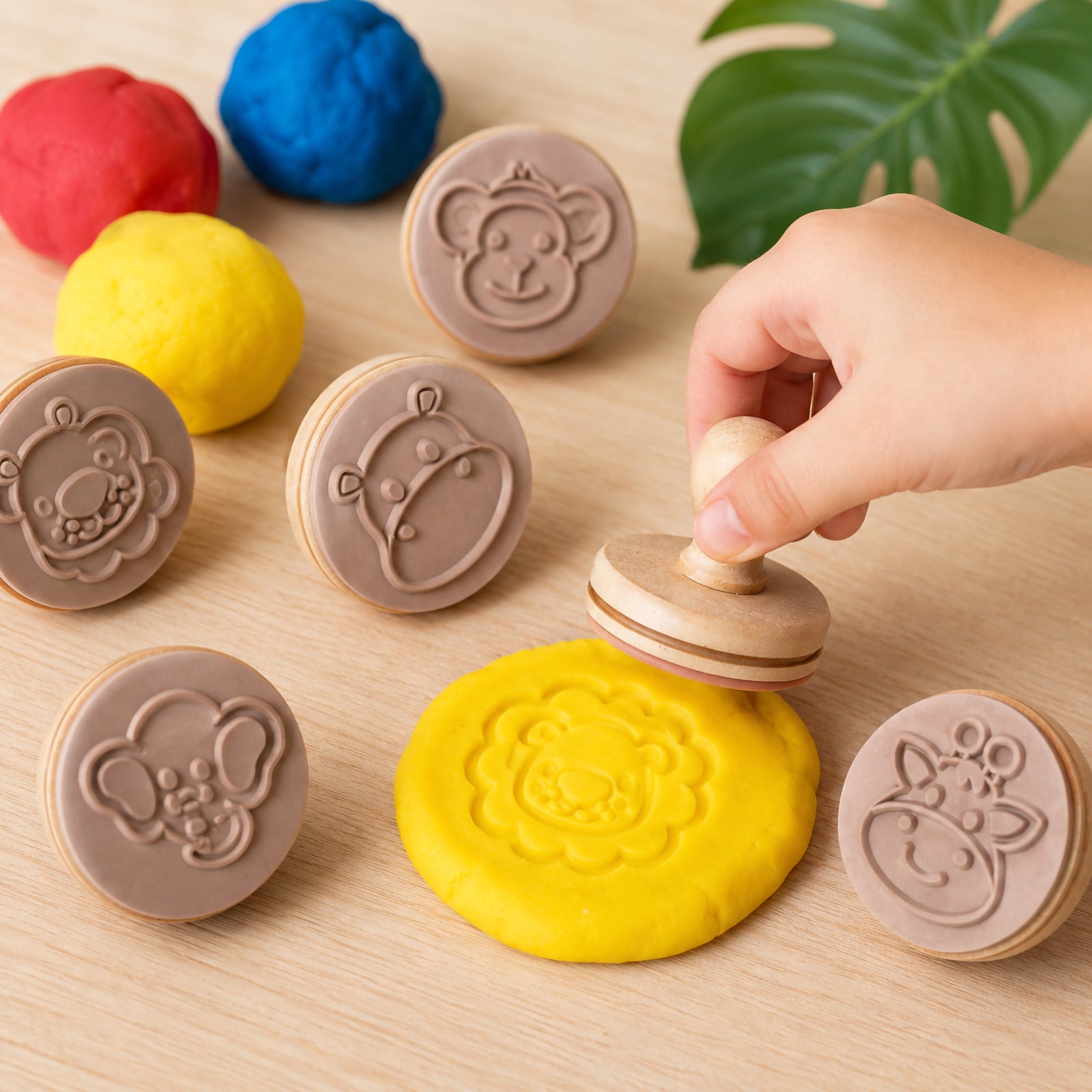 Child using Educational Colours Wooden Dough Stamps Jungle Animals set to press a lion design into yellow dough on a wooden table. The image shows colourful dough balls and jungle themed wooden stamps including monkey, elephant, giraffe and lion designs during sensory play.