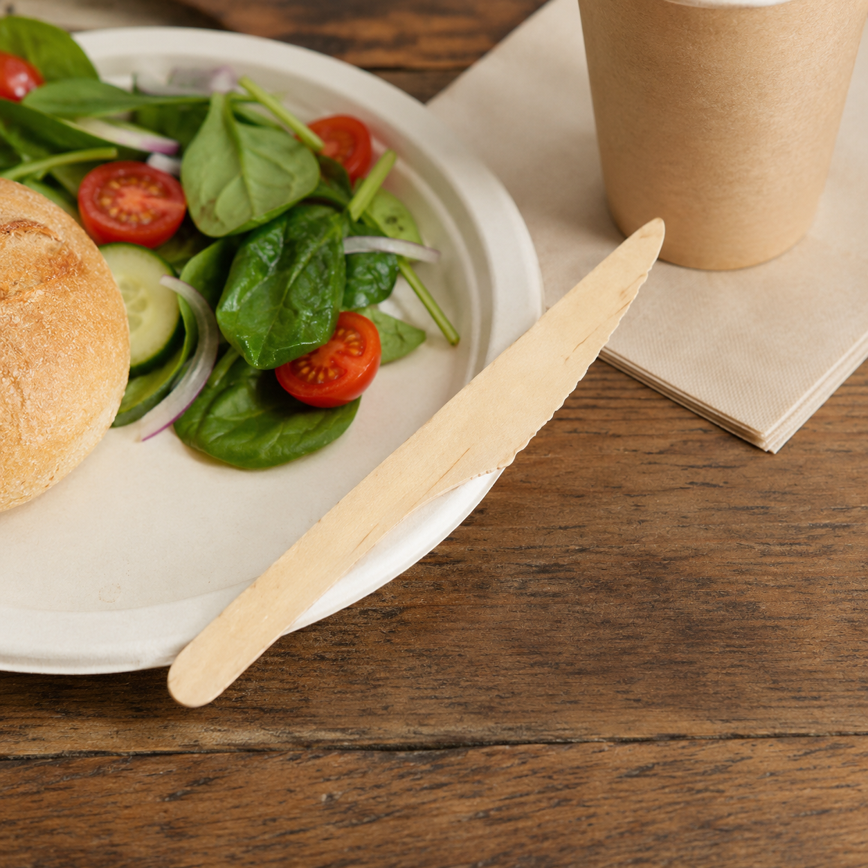Plated salad with a bun and a paper cup on a wooden table, accompanied by a wooden knife.