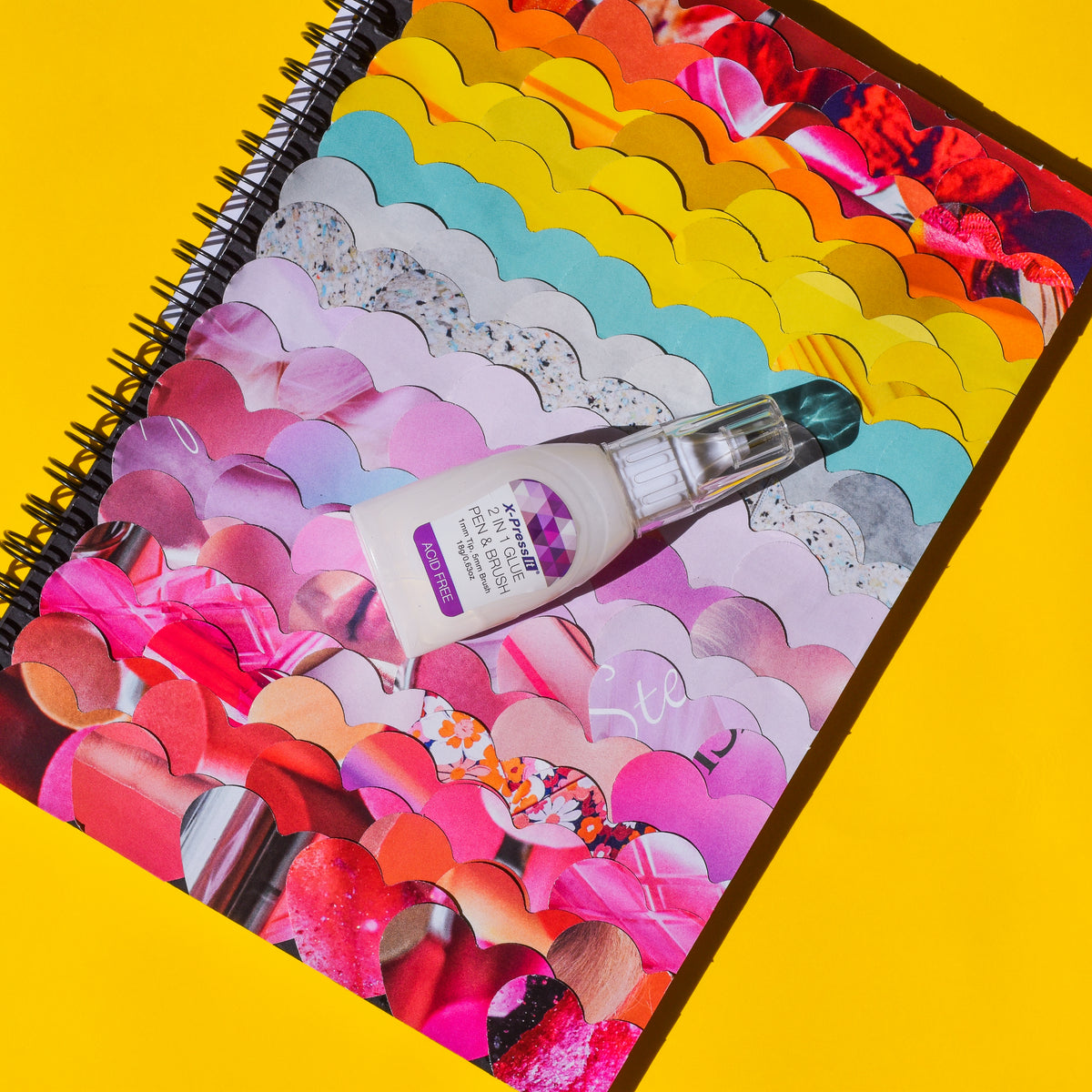 X-Press It 2 in 1 Glue Pen and Brush lying on a spiral notebook covered with a pink and rainbow paper heart collage