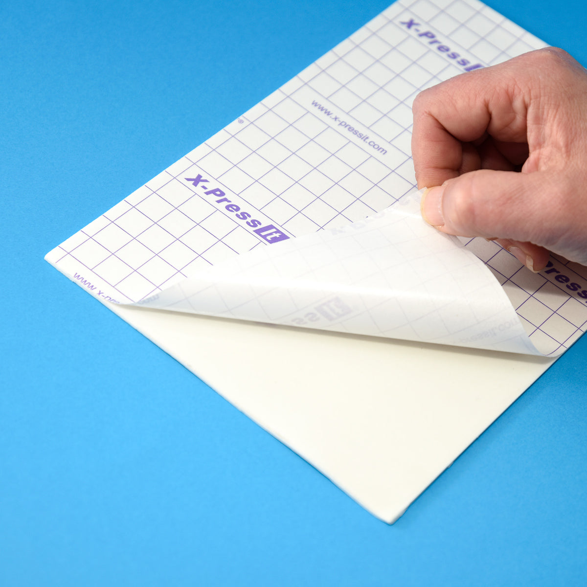 Hand peeling the gridded X-Press It liner back from a white adhesive foam sheet on a blue surface