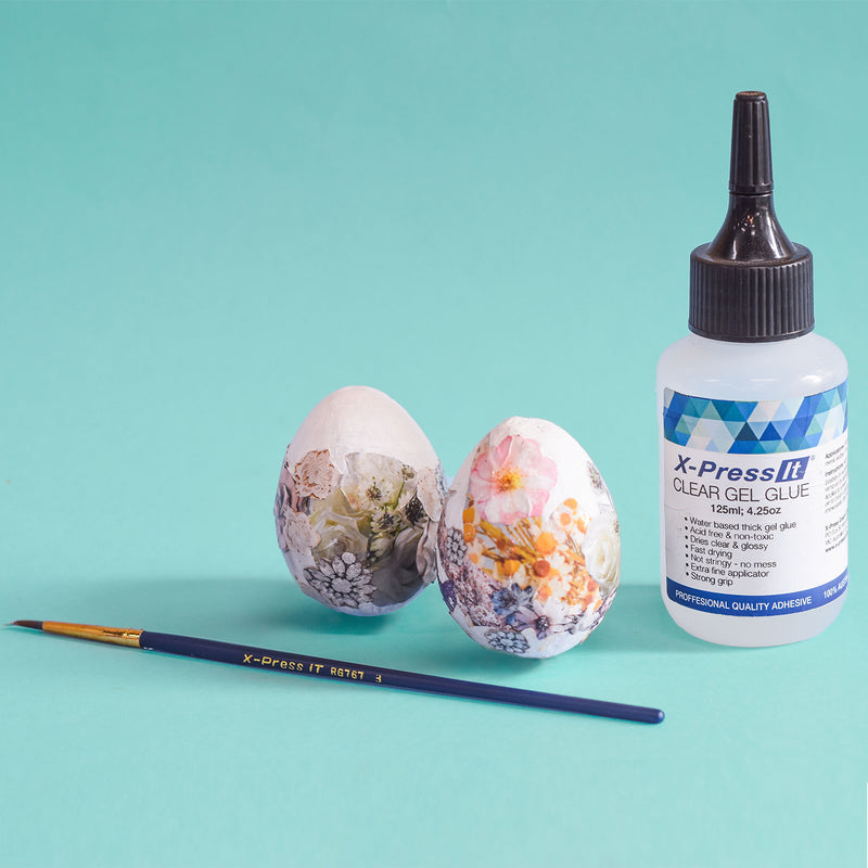 X-Press It Clear Gel Glue 125ml bottle with a black nozzle beside two floral decoupage eggs and a fine paint brush on teal