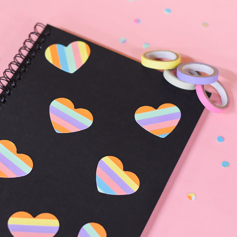 Black spiral notebook decorated with rainbow striped heart shapes cut from deco tape, rolls resting on a pink surface