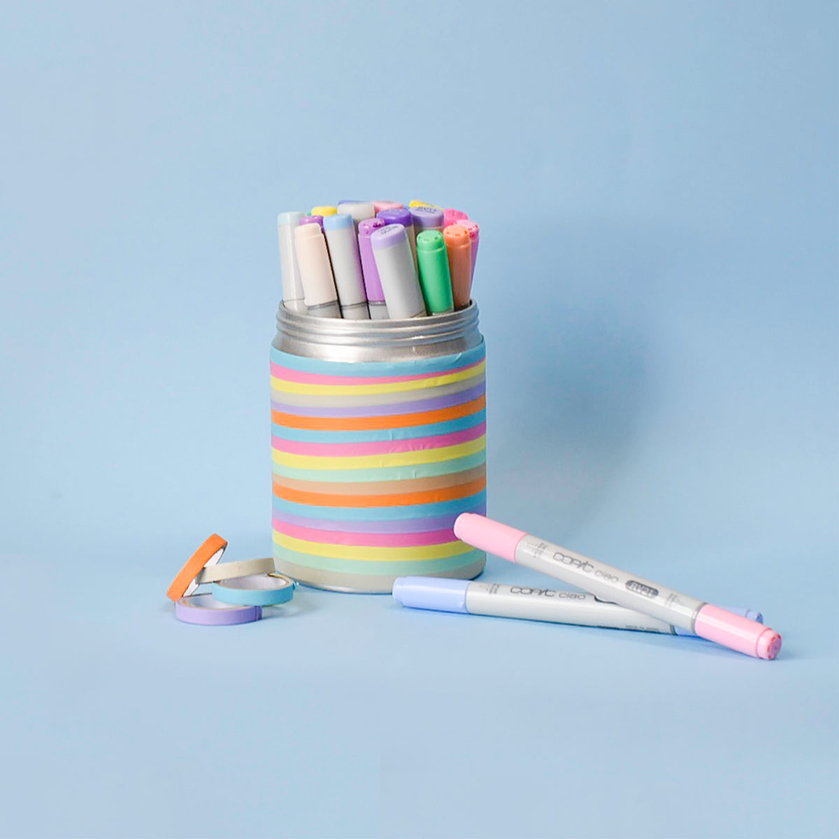 Metal pen pot banded in pastel tape stripes holding marker pens, with loose tape rolls on a lilac background