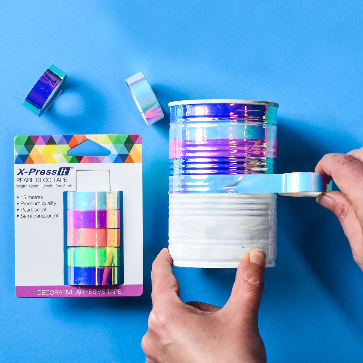 Hand wrapping a tin can with shimmering pearl tape next to the X-Press It Pearl Deco Tape pack on a blue background
