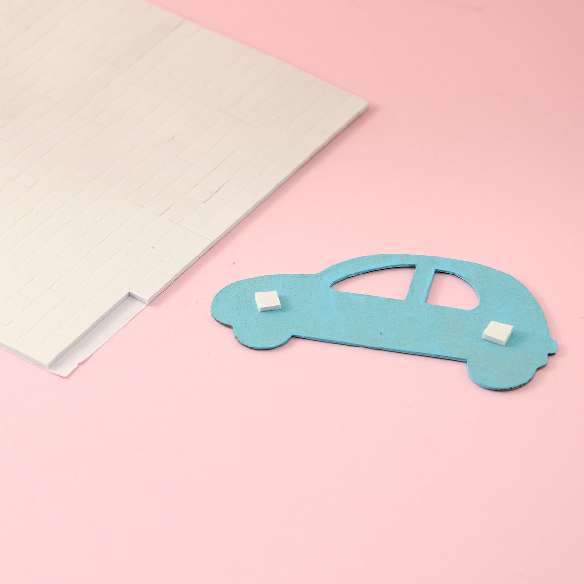 Sheet of white foam squares beside a blue paper car with two squares stuck to its back on a pink background